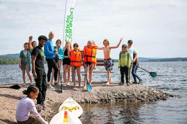 En grupp barn står vid en sjö tillsammans med Carolina Klüft. I förgrunden ser man två SUP:ar och i bakgrunden en Fritidsbanken-flagga. Några barn har flytväst och alla ser glada ut. Foto: Björn Lans