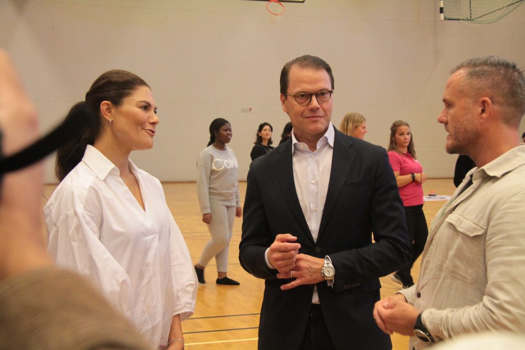 Kronprinsessan Victoria, Prins Daniel och Tino Skovlund, projektledare Fritidsbankens skolsatsning samtalar med varandra i en sporthall. I bakgrunden syns fler personer samtala. Foto: Johanna Grönkvist, Fritidsbanken