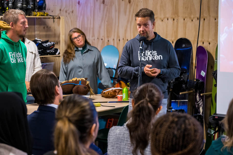 Henric Byström, kommunikationschef på Fritidsbanken Sverige hälsar Kronprinsessparet och övriga gäster välkomna till Fritidsbanken. Foto: PELLE T NILSSON/Swedish Press Agency. På bilden syns Henric Byström vid en mikrofon, tillsammans med Carina Haak och David Mathiasson. I förgrunden ser man bland annat Kronprinsessparet bakifrån. I bakgrunden står några snowboards lutade mot en vägg.